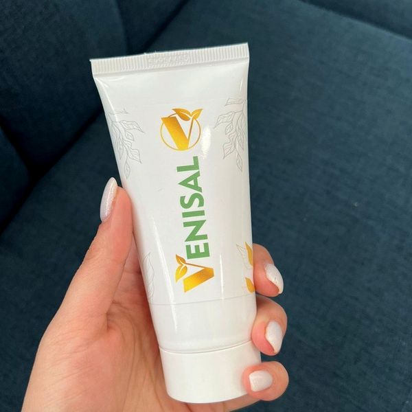 Roka drži belo tubo kreme Venisal z zlatim logotipom pred modro sedežno garnituro